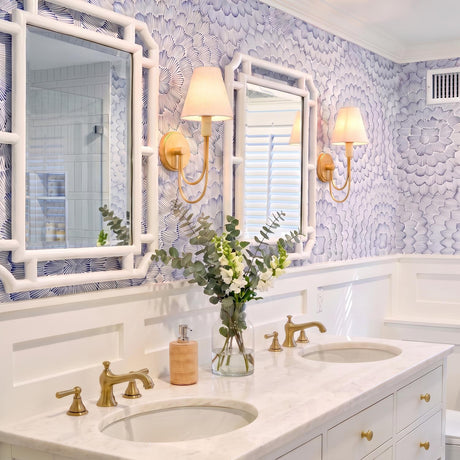 A coastal bathroom features gold leaf wall sconces with linen shades mounted against blue patterned wallpaper, flanking white bamboo mirrors above a marble double vanity.