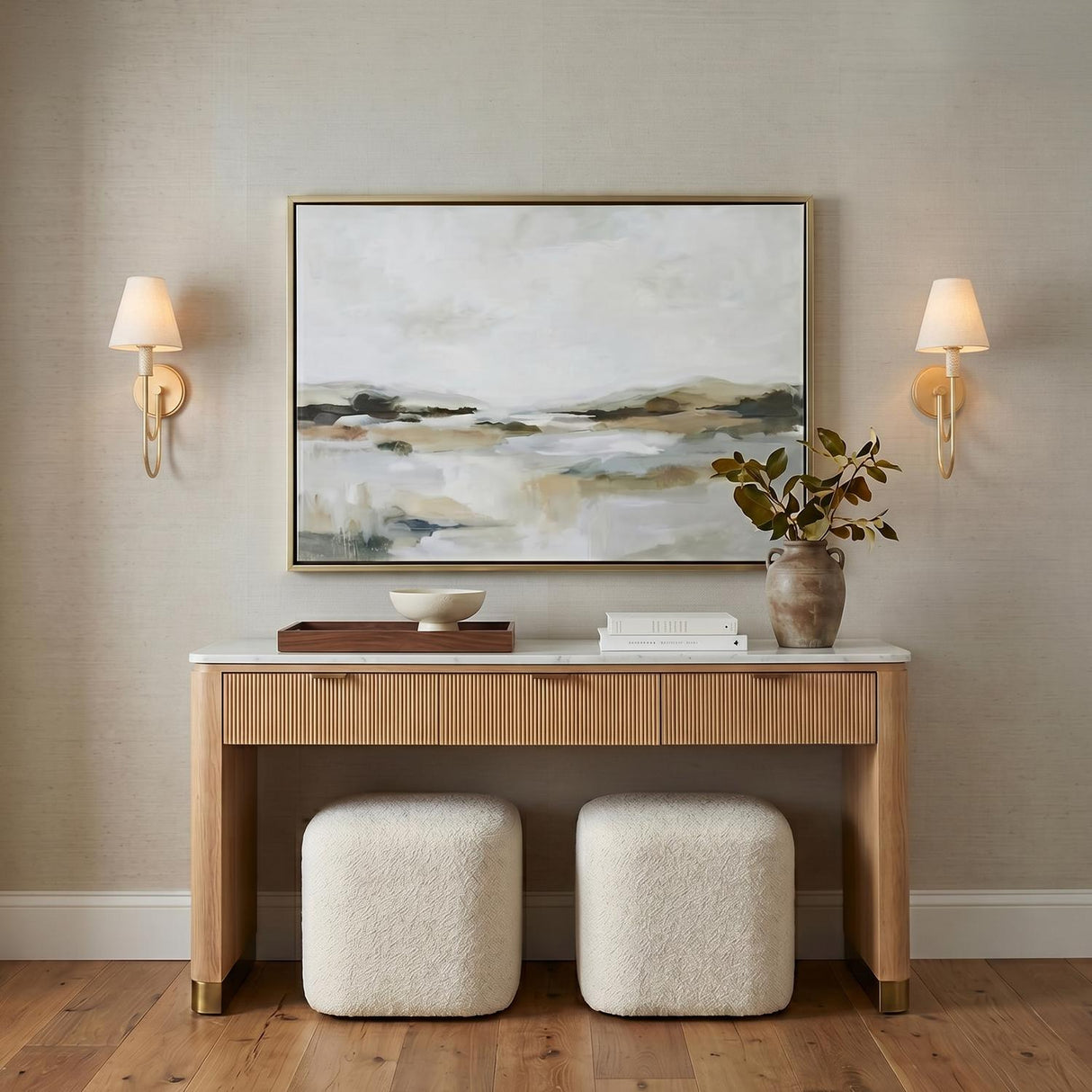 A transitional foyer with two gold leaf wall sconces with white linen shades flanking a landscape painting above a fluted wood console table with a marble top.