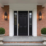 A traditional colonial red brick entry with two matte black and patina brass wall lanterns flanking a black front door at twilight, featuring stone steps and boxwood.