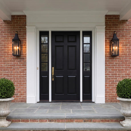 A traditional colonial red brick entry with two matte black and patina brass wall lanterns flanking a black front door at twilight, featuring stone steps and boxwood.
