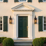 A colonial home exterior with two matte black and patina brass wall lanterns flanking a dark green front door on white stucco at dusk with boxwood shrubs.