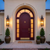 A Mediterranean entry with two black and patina brass Dante Outdoor Wall Lights flanking a burgundy arched door at twilight, mounted on cream stucco with potted evergreens.