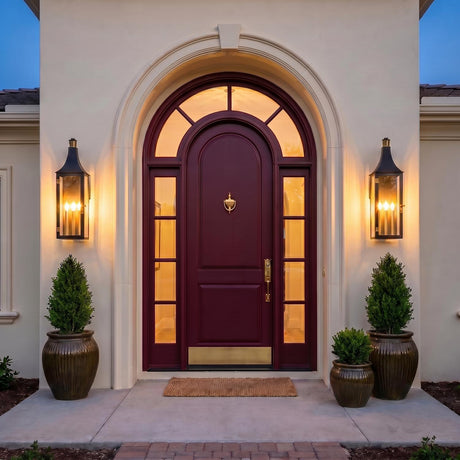 A Mediterranean entry with two black and patina brass Dante Outdoor Wall Lights flanking a burgundy arched door at twilight, mounted on cream stucco with potted evergreens.