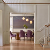 A transitional dining room with the Orly 4 Light Linear Chandelier in patina brass and seeded cloud glass under a double-height ceiling with purple chairs.