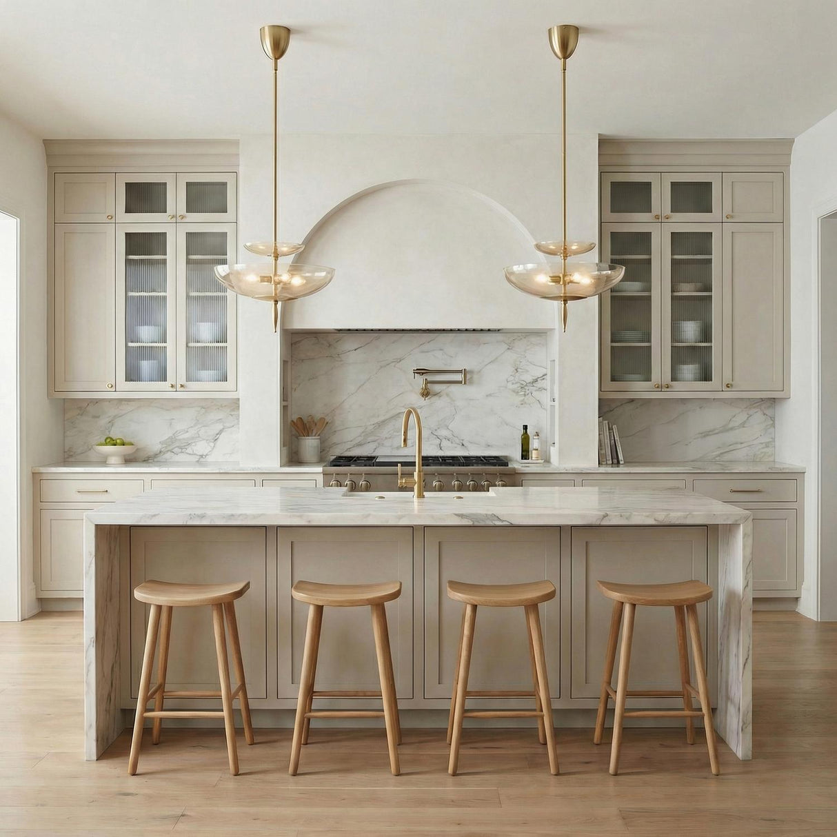 A contemporary kitchen features two Zara Large Two-Tier Pendant 21", Brushed Brass and Chamomile Glass fixtures over a white marble waterfall island and four wooden stools.