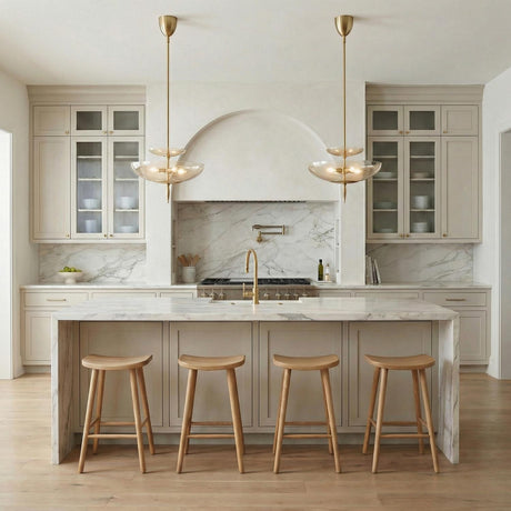 A contemporary kitchen features two Zara Large Two-Tier Pendant 21", Brushed Brass and Chamomile Glass fixtures over a white marble waterfall island and four wooden stools.