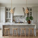 A transitional kitchen features two Zara Large Three-Tier Pendants in brushed brass and chamomile glass hanging over a marble waterfall island and heavily veined backsplash.