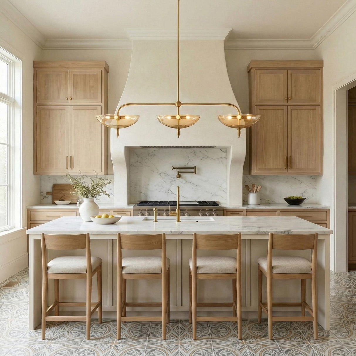 A transitional kitchen features the Zara Linear Pendant 54-inch in Brushed Brass and Chamomile Glass suspended over a white marble waterfall island with four light wood stools.