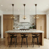A transitional kitchen features three Zara Medium Two-Tier Pendant 14-inch, Bronze and Chamomile Glass pendants over a white marble island with light wood cabinets and dark stools.