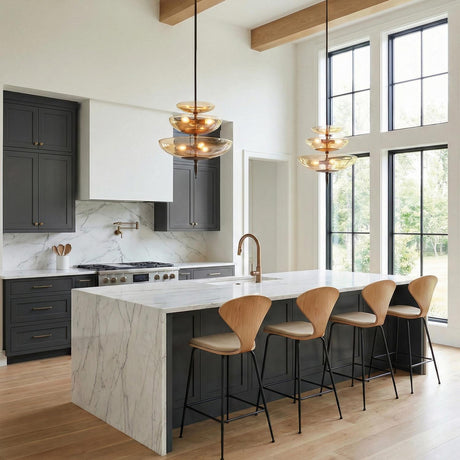 A contemporary kitchen with two Zara Large Three-Tier Pendants in bronze and chamomile glass over a white marble waterfall island and dark cabinets under a beamed ceiling.