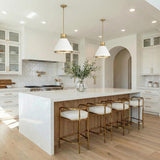 A contemporary kitchen features the Drew Medium Pendant 19 in matte white and aged brass over a white quartz waterfall island with wood fluting and brass barstools.
