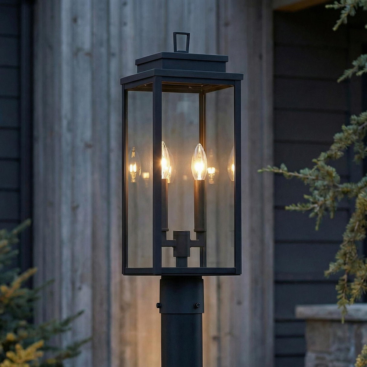 An outdoor entryway featuring the Trevor Outdoor Post Light 20", Black with clear glass panes positioned near a house with dark siding and evergreen trees.