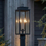 An outdoor entryway featuring the Trevor Outdoor Post Light 20", Black with clear glass panes positioned near a house with dark siding and evergreen trees.