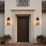A Mediterranean entryway featuring two matte black Trevor Outdoor Wall Light lanterns flanking a dark wood front door set within a stone surround and brick pathway.