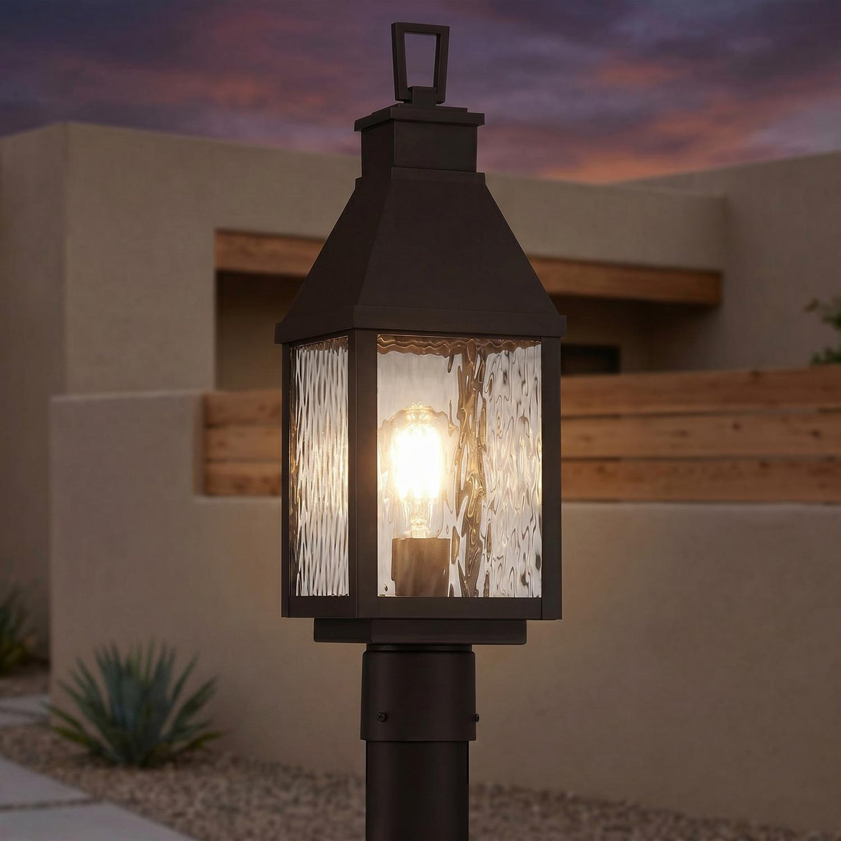 A Mediterranean exterior featuring the Westin Outdoor Post Light 20", Bronze and Water Glass glowing at twilight on a post beside beige stucco walls and agave.