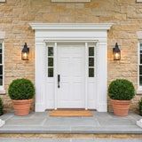 A traditional stone home exterior featuring two matte black Westin Outdoor Wall Light fixtures flanking a white front door, with boxwood shrubs in terracotta pots at dusk.