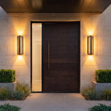 A modern home entry with two Zenith Outdoor LED Wall Light, Large 24", Bronze fixtures flanking a dark wood door on stone walls at night with shrubs.
