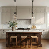 A transitional kitchen featuring two Naya Medium Pendant 12", Matte Black and Clear Glass fixtures over a marble-topped walnut island with beige cabinets and marble backsplash.