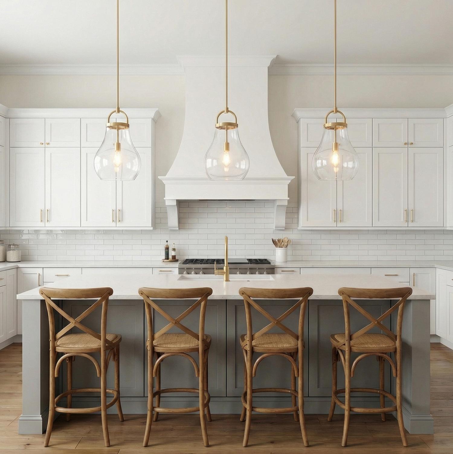 A transitional kitchen features three Naya Medium Pendant 12 inch in Satin Brass and Clear Glass suspended over a white marble island with gray cabinetry and wooden stools.
