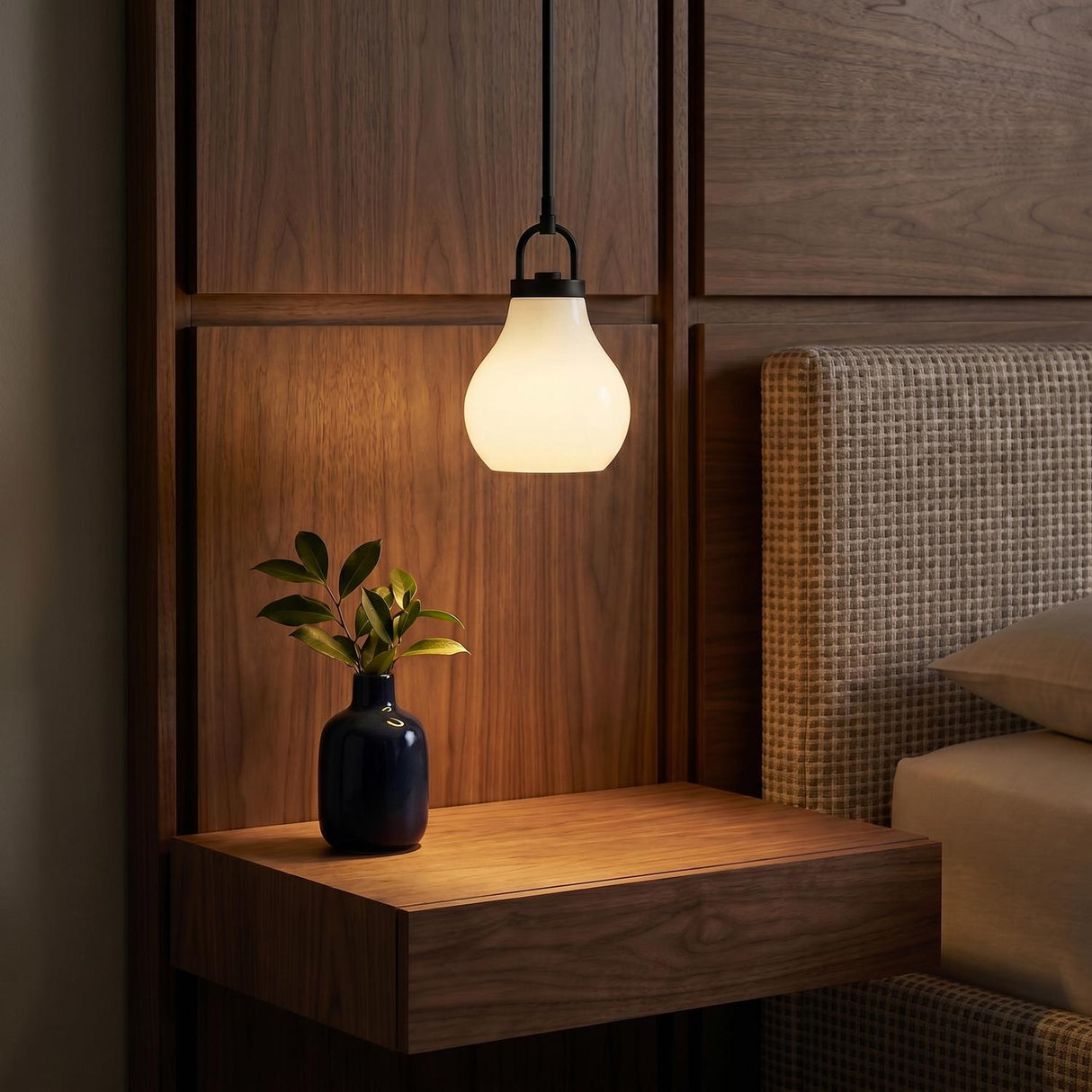 A mid-century bedroom with a matte black and opal glass pendant suspended over a walnut floating nightstand against walnut wood wall paneling and a textured headboard.