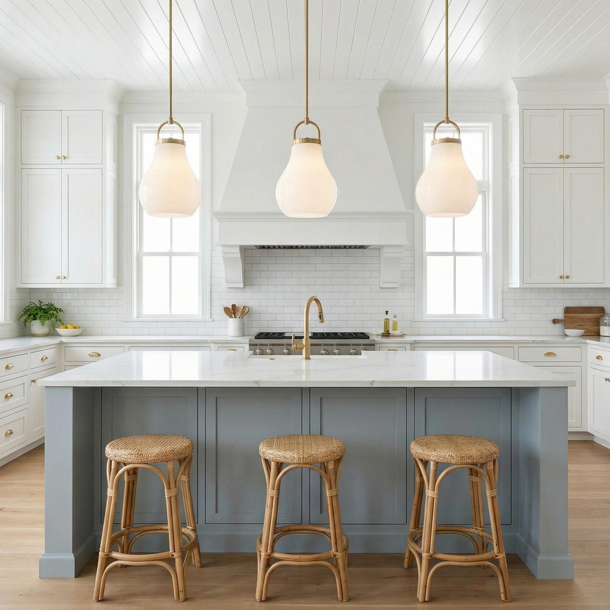 A farmhouse kitchen featuring three Naya Medium Pendant 12", Satin Brass and Opal Glass fixtures over a marble island with blue-gray cabinets and woven rattan stools.