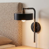 A modern bedroom features an Amadeo Wall Sconce 18", Black and Bronze mounted on a white wall beside a patterned upholstered headboard and light wood nightstand.