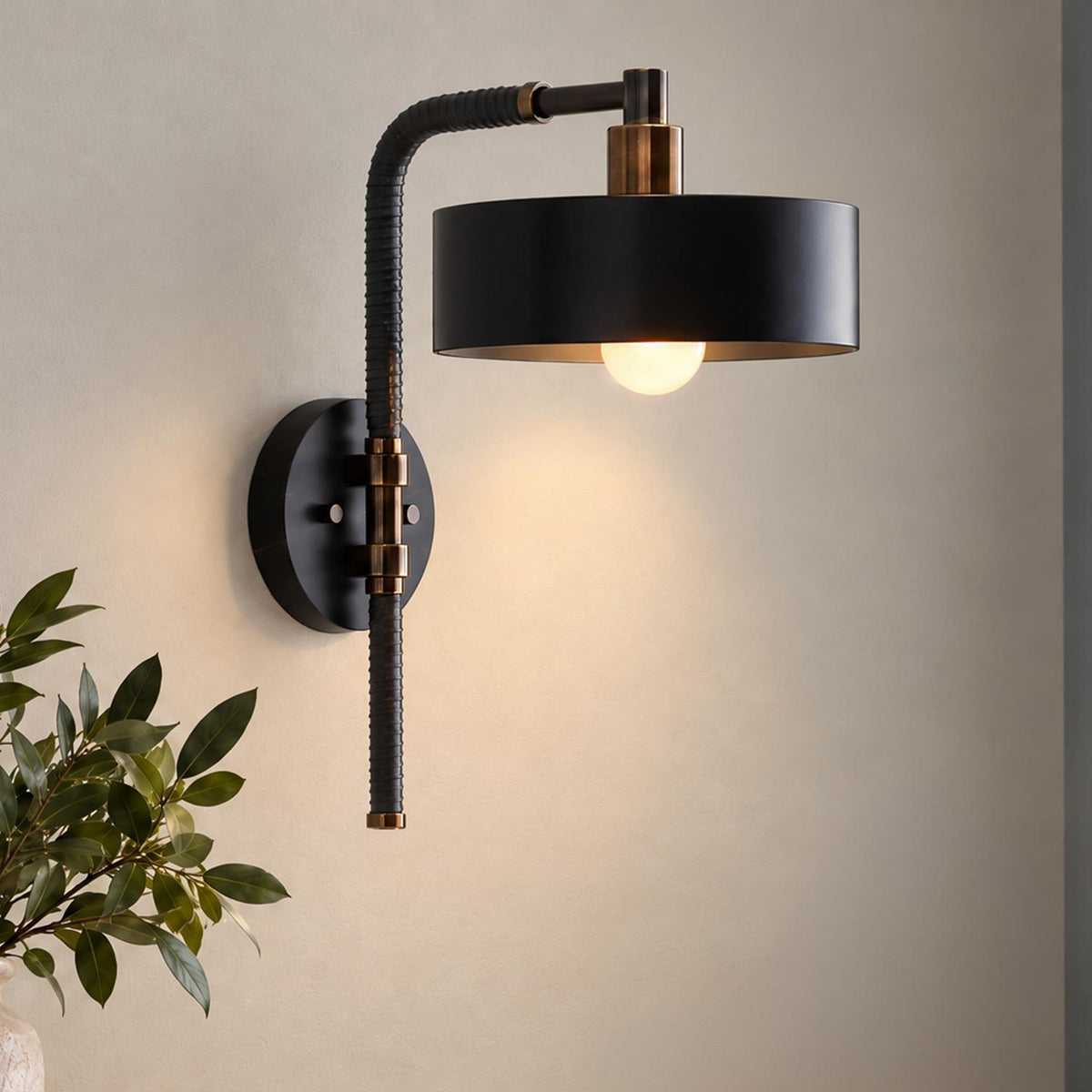 A modern living room featuring an Amadeo Wall Sconce in black and bronze mounted on a neutral wall beside a leafy green potted plant.