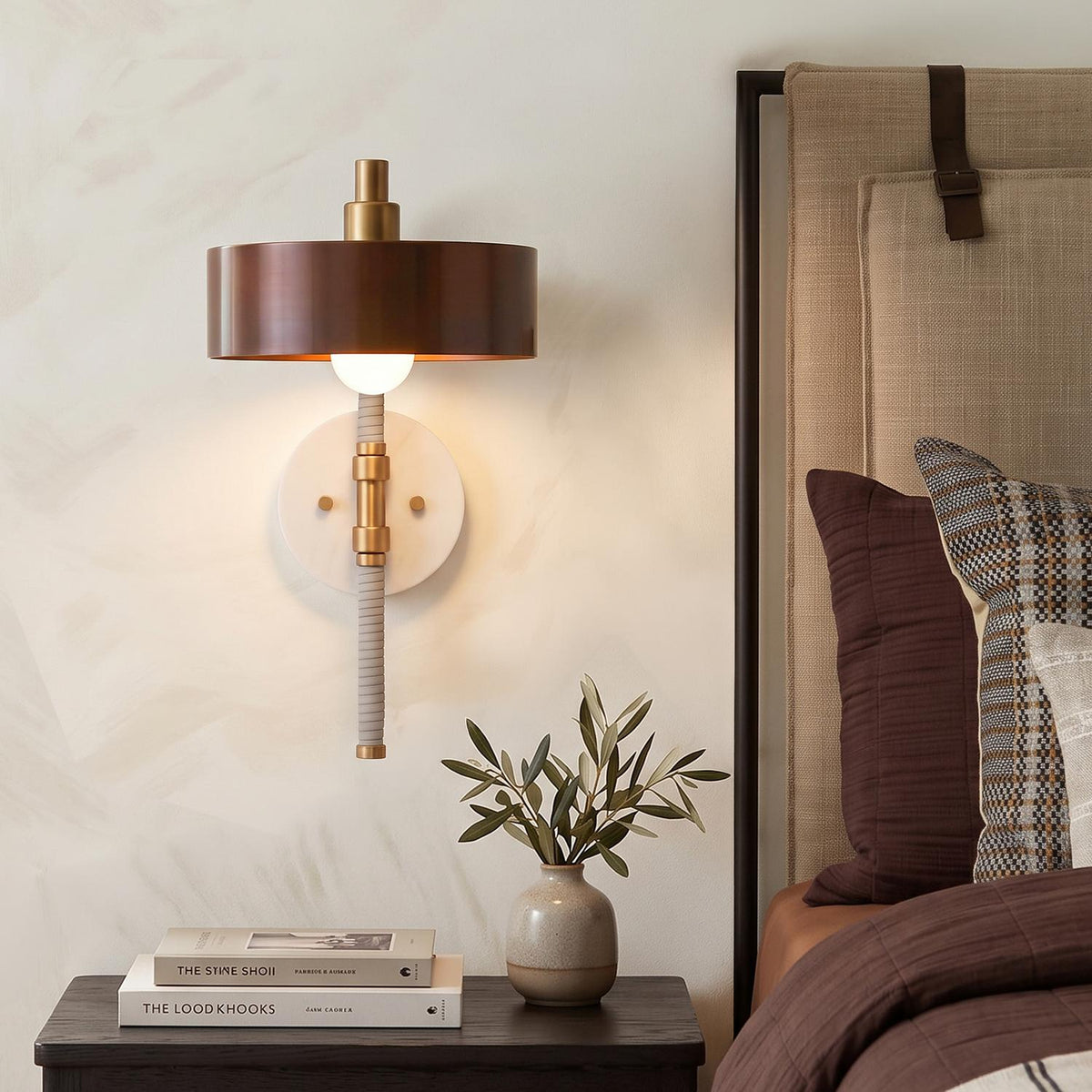 A transitional bedroom with a heritage brass and brown wall sconce mounted above a dark wood nightstand beside a neutral upholstered headboard and olive plant.