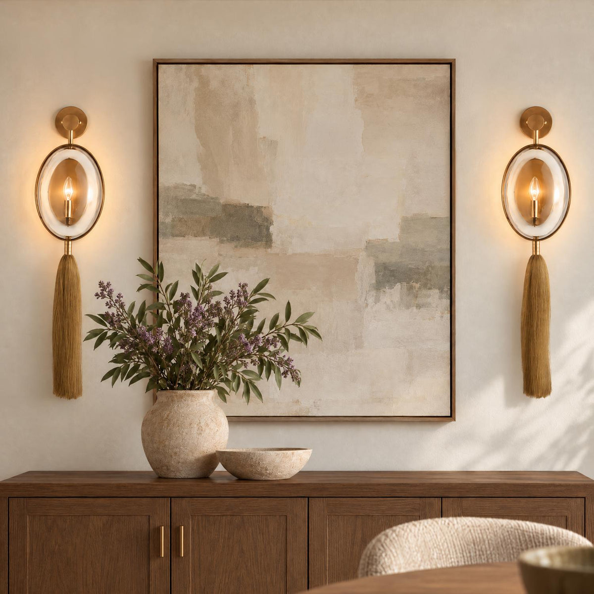 A transitional dining room featuring two antique brass wall sconces with glass accents and tassels flanking framed abstract art above a dark wood credenza.