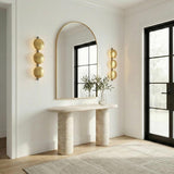 A contemporary entryway featuring two Ilyana Wall Sconce 32 in antique brass flanking an arched mirror above a travertine console table and black framed door.