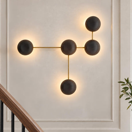 A modern foyer displays the Hanley 5 Light Sculptural Wall Sconce with matte black ceramic spheres and brass accents on a paneled wall beside a wooden staircase.
