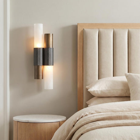 A modern bedroom featuring the Rainier Wall Sconce in brushed bronze with charcoal leather accents mounted beside a beige channeled headboard and a marble topped nightstand.