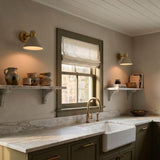 A modern farmhouse kitchen with the Cosette Adjustable Wall Sconce in soft taupe and antique brass flanking a window over a marble countertop and olive green cabinets.