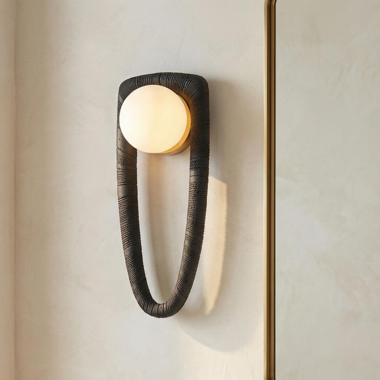 An entryway featuring the Iliana Wall Sconce in charcoal stone and opal glass mounted on a textured plaster wall next to a gold-framed mirror.