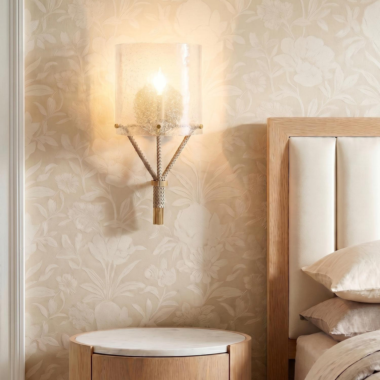 A transitional bedroom with the Nadine Wall Sconce featuring clear seeded glass and taupe leather accents mounted on floral wallpaper above a marble-topped wood nightstand.