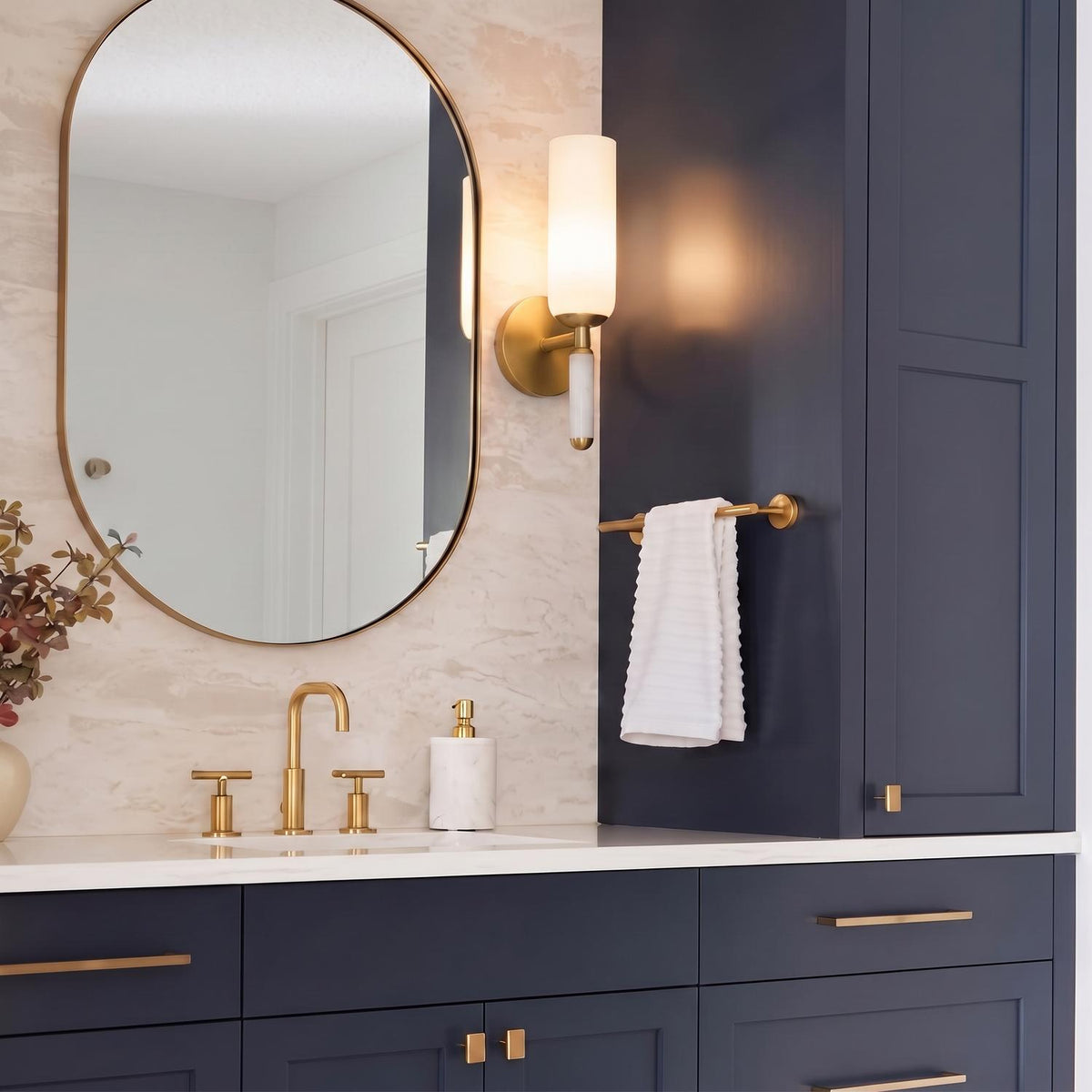 A transitional bathroom featuring the Riona Wall Sconce 15", Antique Brass and Opal Glass mounted beside a navy cabinet above a white marble backsplash and gold faucet.