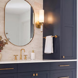 A transitional bathroom featuring the Riona Wall Sconce 15", Antique Brass and Opal Glass mounted beside a navy cabinet above a white marble backsplash and gold faucet.