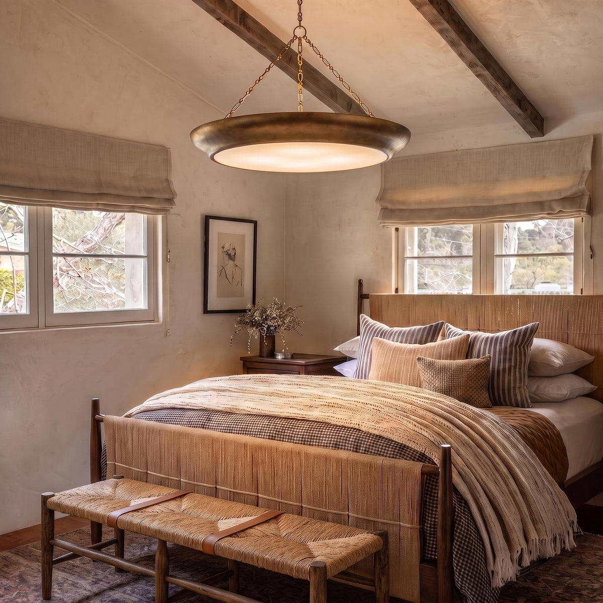 A transitional bedroom with exposed wooden ceiling beams features a large antique brass saucer pendant suspended over a woven bed dressed in layered neutral linens.