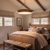 A transitional bedroom with exposed wooden ceiling beams features a large antique brass saucer pendant suspended over a woven bed dressed in layered neutral linens.