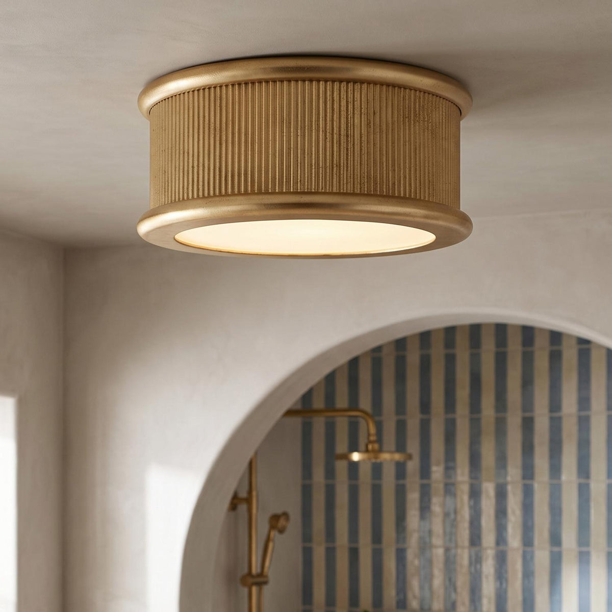 A contemporary bathroom features a gilded brass flush mount ceiling light mounted near an arched shower entry with blue and beige striped tile.