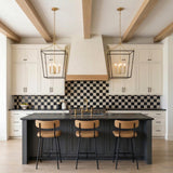 A transitional kitchen features two Cambria Extra Large Lantern Pendant 24", Matte Black and Modern Brass over a dark island with a checkered tile backsplash and wood beams.