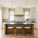A transitional kitchen features two Cambria Extra Large Lantern Pendant 24", Matte Black and Modern Brass fixtures over a wood island with black countertops and cream cabinets.