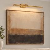 A modern living room features the Bria LED Picture Light 24", Modern Brass mounted above framed abstract artwork with a textured ceramic vase.