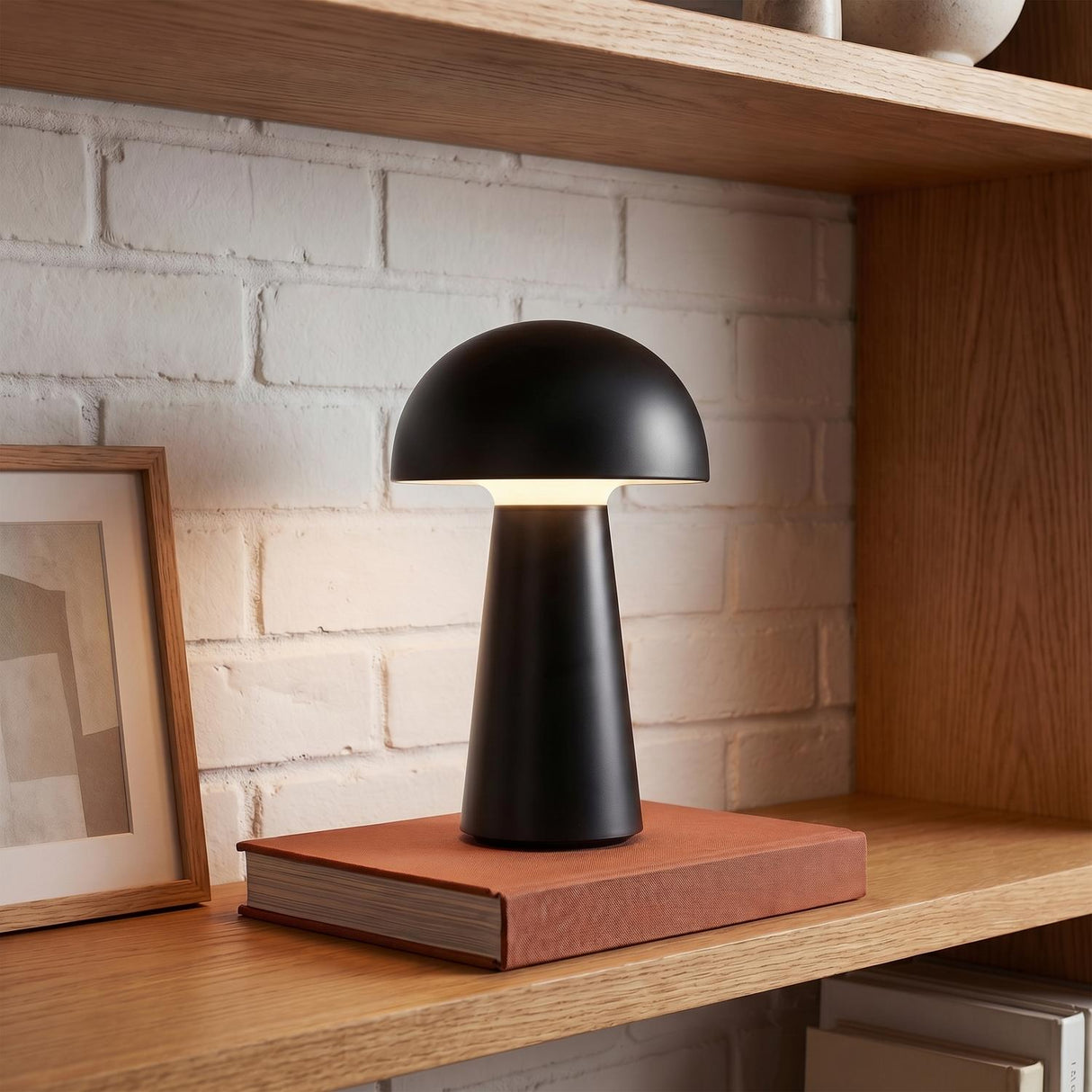 A modern living room featuring the matte black Marnie Rechargeable Table Lamp 9" resting on an orange book atop a light oak shelf with white brick.