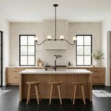 A contemporary kitchen features the Leora 4 Light Linear Pendant 48", Matte Black with frosted glass globes over a white marble island and fluted wood cabinets.