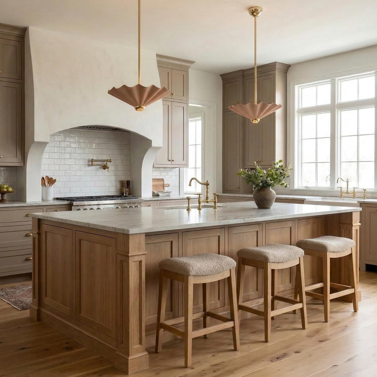 A transitional kitchen features two Babette Medium LED Pendant 16" fixtures in blush and brass over a white marble island with three wooden stools and taupe cabinetry.