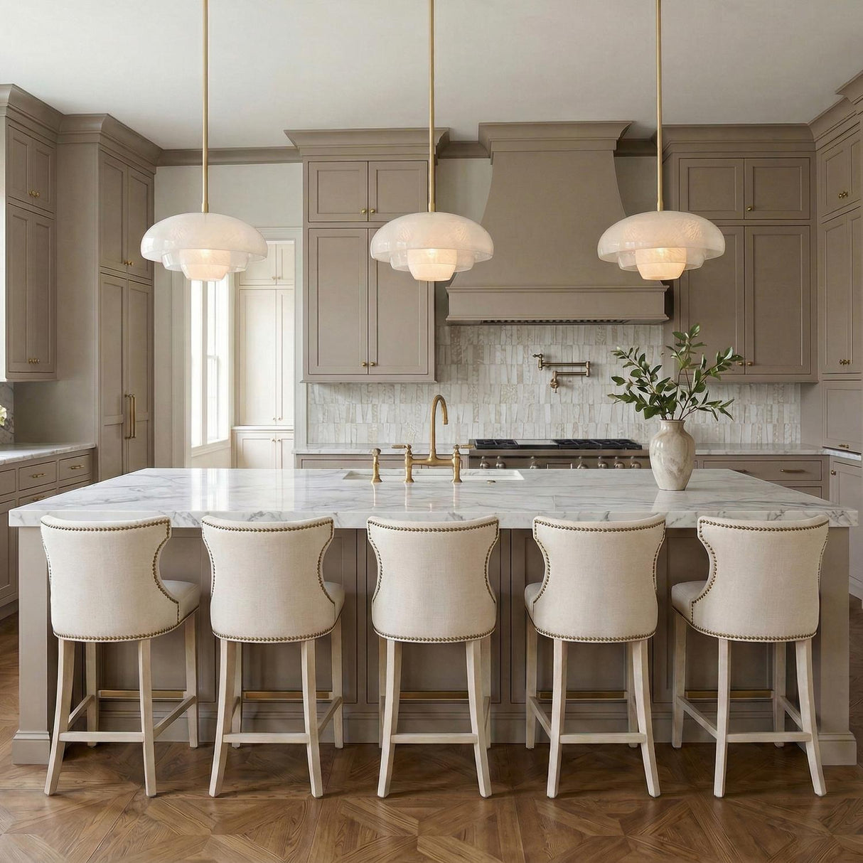 A transitional kitchen features three Jalina Pendant 18", Aged Brass and Art Glass fixtures over a white marble island with taupe cabinets and five upholstered bar stools.