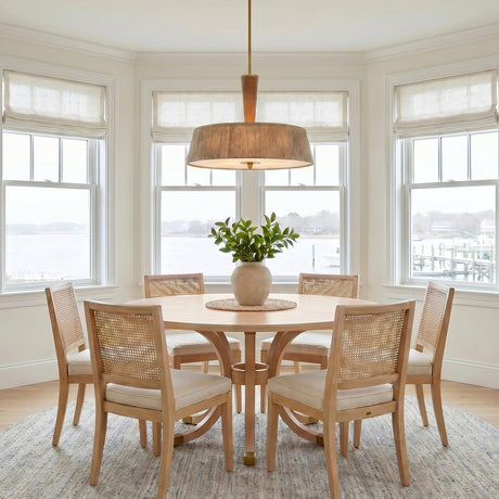 A coastal dining room with a jute and aged brass drum pendant over a round light wood table and rattan chairs set within a bay window.