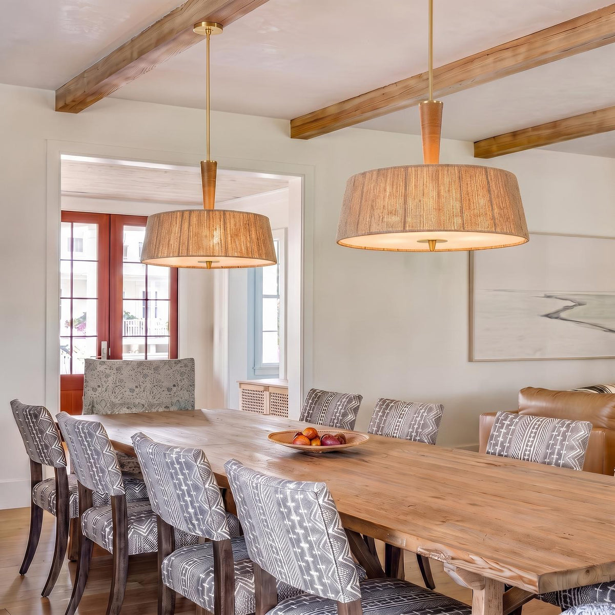 A coastal dining room with two jute and aged brass pendants suspended over a long natural wood table under a beamed ceiling with pattern upholstered chairs.