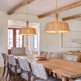 A coastal dining room with two jute and aged brass pendants suspended over a long natural wood table under a beamed ceiling with pattern upholstered chairs.